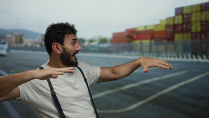 Fototapeta premium Hispanic man with beard gesturing playfully at a port with ships and containers in the background, creating a lively scene with maritime and industrial vibes.
