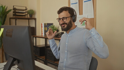 Hispanic man with beard wearing headset in office, expressing happiness during a video call in a professional environment featuring a computer and office supplies visible.