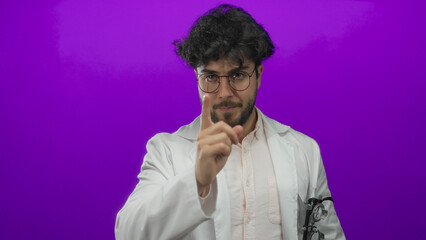 Obraz premium Young man with beard and glasses in white coat standing against a vibrant purple background displaying a serious expression in a studio setting.