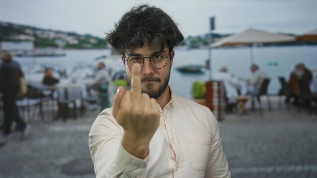Young hispanic man with beard making rude gesture on a terrace at an outdoor coffee restaurant.