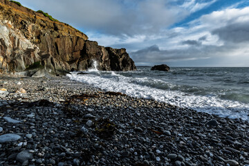 Coastal scene of waves hitting a rocky beach on a cloudy day. A cliff dominates the horizon.