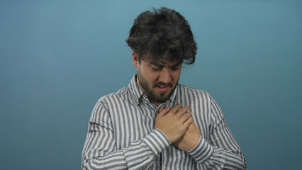 Young hispanic man with beard clutching chest against blue background in a moment of pain, suggesting a heart condition or emotional distress.