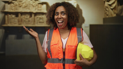 Woman wearing orange safety vest holds yellow hardhat and raises hand palm up in building; excitement career growth.