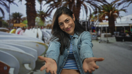 Fototapeta premium Woman with hands crossed over chest wearing denim jacket and striped crop top on street with surfboards and palm trees; gratitude connection.
