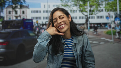 Obraz premium Woman making phone hand gesture to ear on a busy city street in denim jacket and striped shirt; joy connection playfulness.