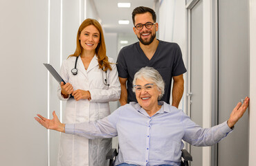 Obraz premium Portrait of confident doctors pulling cheerful senior patient sitting in wheelchair along hospital corridor