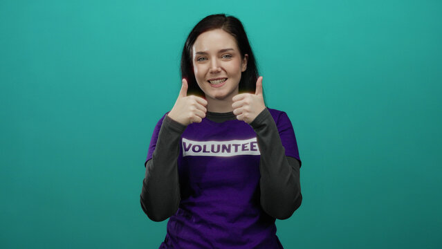 Brunette woman wearing purple volunteer shirt against green background showing thumbs up in support of charity and community service projects.