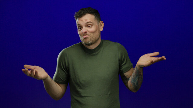 Young man with tattooed arm in green shirt shrugging and expressing confusion against blue background