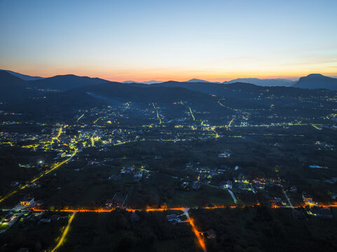Aerial view of a tranquil town nestled amidst rolling hills, illuminated by the warm glow of streetlights against the deepening twilight, Santo Stefano del Sole, Irpinia, Campania, Italy.
