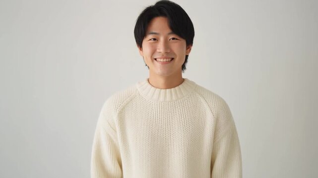20代の日本人男性が、クリーム色のニットとベージュのチノパンでやわらかく微笑む冬のスタジオシーン。
白背景と温かな光が、ナチュラルな黒髪ときれいな肌質を際立たせ、親しみやすい雰囲気を演出。
