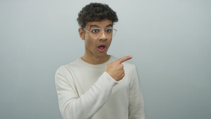 Young hispanic man points finger to the right in studio with pale blue wall and open mouth; surprise.