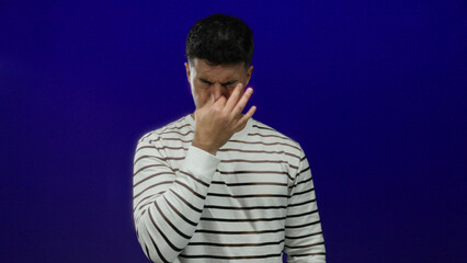 Obraz premium Young man pinching nose in disgust, isolated against blue wall, wearing striped shirt, expressing distaste, adult male reacting to unpleasant odor, over blue background