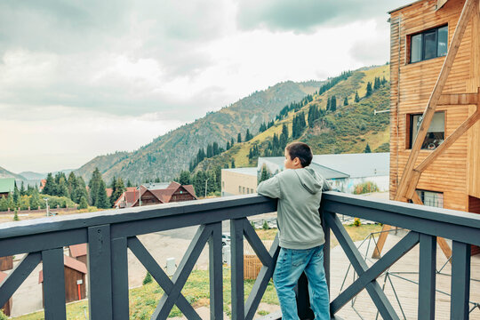 young Asian man stands on a wooden balcony overlooking green mountains. The scene captures a tranquil moment in a recreational travel setting. - Powered by Adobe