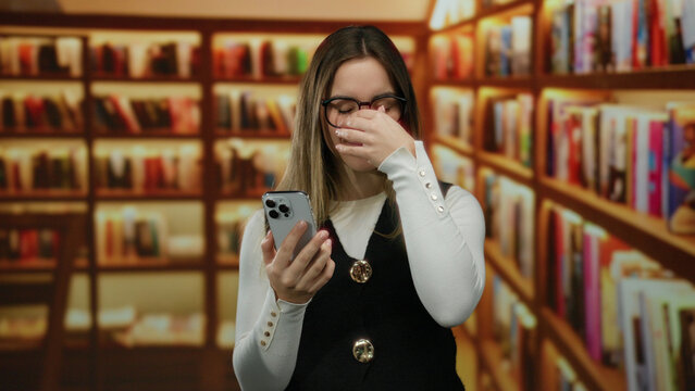 Woman holding smartphone in library with blonde hair and eyeglasses, showcasing a reflective mood under warm lighting among rows of bookshelves indoors.