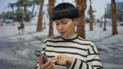 Woman with short hair in city street outdoor setting using smartphone focuses on device with palm trees in blurred background