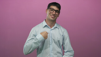 Man wearing glasses in light blue shirt smiling and pointing finger to chest against pink studio wall; confidence.