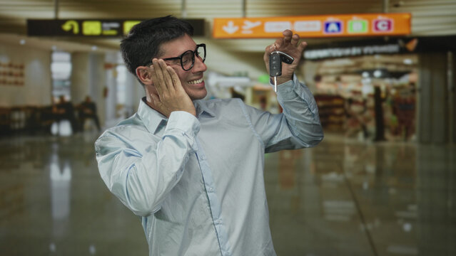 Man smiling broadly while holding car key fob at airport terminal and touching his cheek; excitement.