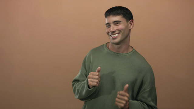Man smiling and dancing with raised hands and thumbs up gesture in studio wearing green sweater; pure joy.