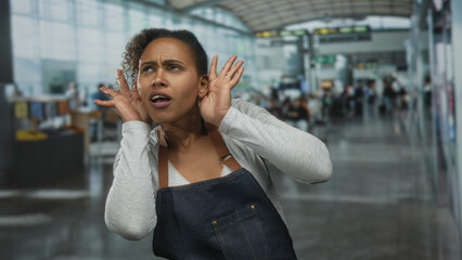 Fototapeta premium Woman cups both hands to ear and squints while listening in a bustling airport terminal wearing denim apron and long sleeve top; frustration seeking help.
