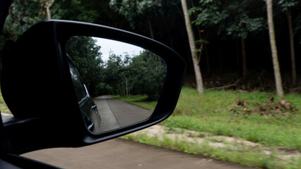 Mirror view of car on the concrete road. Side of road with deep forest. Reflection in the mirror consists of the side of a car and a road with forests on both sides.