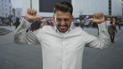 Hispanic man grinning widely and pointing thumbs to chest on a bustling downtown street; self...