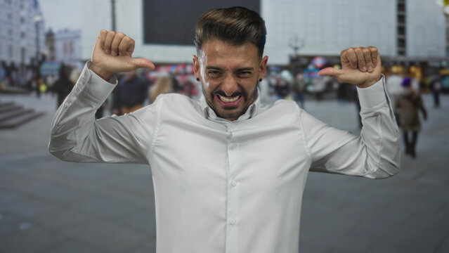 Hispanic man grinning widely and pointing thumbs to chest on a bustling downtown street; self confidence.