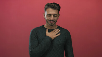 Obraz premium Young hispanic man with hand on chest standing in studio against red wall wearing dark knit sweater; sincerity.