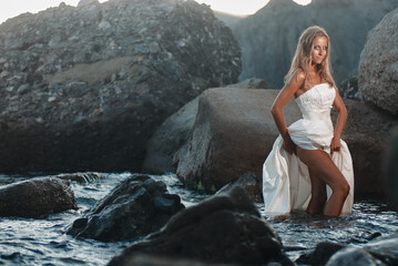 Bride in White Dress Poses on Coastal Rocks During Sunset Beach Photoshoot