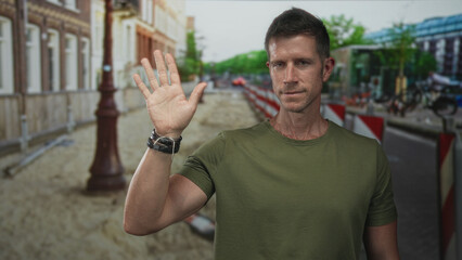 Man in green t shirt raises open hand in a wave on a city street near construction barriers; greeting warmth.