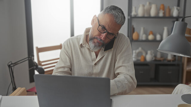 Man with glasses and gray beard holding phone to ear while typing on laptop at desk in a building office workspace; stress deadline.