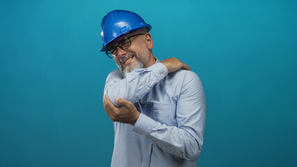 Man architect wearing blue hardhat and glasses smiling while holding elbow in teal studio; confidence safety planning.