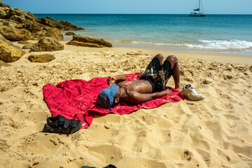 Young man sunbathing on red blanket on secluded Figueira Beach by calm tranquil ocean water idyllic Algarve Portugal