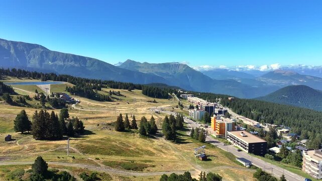 Aerial drone footage of a mountain road, ski lift, and residential buildings in Chamrousse, French Alps, during summer. The scene captures green slopes, pine forests, and panoramic alpine peaks under 
