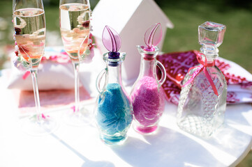 Bright outdoor table with champagne flutes, blue and pink sand-filled bottles, and crystal decanter...
