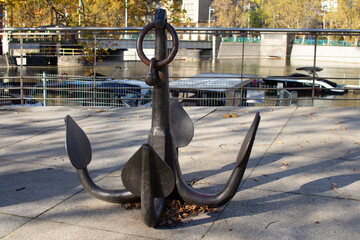 Large metal anchor displayed near a river in a city park during autumn