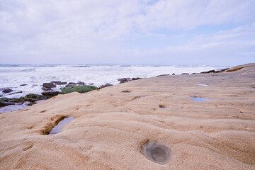 A sandy beach with small puddles and rocks near the ocean under a cloudy sky.