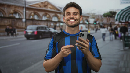 Hispanic man points finger to smartphone while holding phone on busy city street; happiness connection sharing.