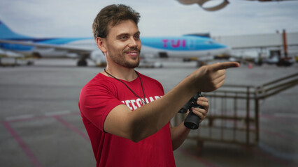 Lifeguard man smiling pointing finger at airport tarmac wearing red shirt and holding binoculars;...
