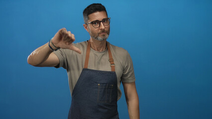 Fototapeta premium Man showing thumbs down gesture wearing denim apron in blue studio; disapproval critique feedback review.