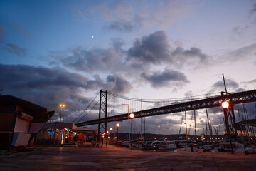 A scenic view of a suspension bridge at dusk with a marina in the foreground. The sky is partly cloudy with a crescent moon visible. Street lamps illuminate the area, creating a warm ambiance.