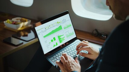 Unrecognizable businessman working on his laptop computer, analyzing financial charts and graphs while flying on a luxurious private jet during a business trip to a new destination - Powered by Adobe