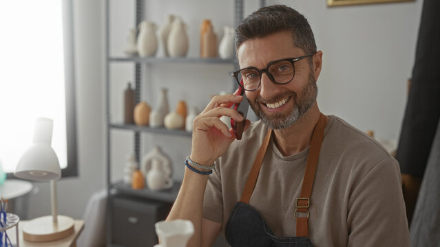 Man holding smartphone to ear, smiling in studio with pottery on shelves while wearing apron and glasses; creative connection joy.