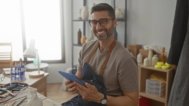 Man smiling, holding a blue tablet with hands visible and wearing an apron in a studio surrounded by pottery and tools; contentment creativity craft.
