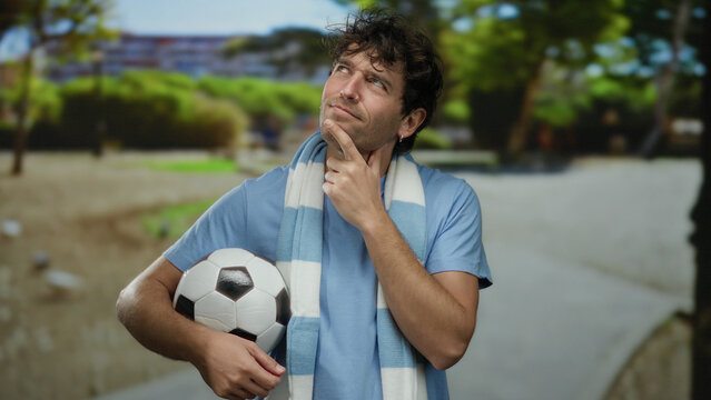 Hispanic man in park holds soccer ball, contemplating thoughtfully, surrounded by green scenery and showcasing athletic appeal in a sporty, outdoor setting.