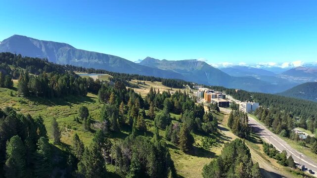 A stunning aerial drone view of the Chamrousse ski resort in the French Alps during summer. The footage shows a scenic mountain landscape with a clear blue lake, evergreen forests