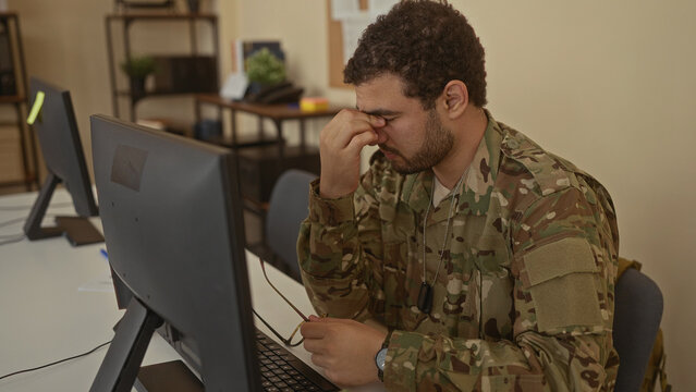 Soldier in camouflage uniform pinches nose while seated at computer desk in office workplace; disgust.