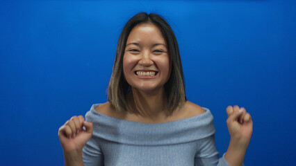 Fototapeta premium Chinese woman smiling and expressing joy against a vivid blue wall background with arms raised showcasing excitement and happiness in a studio setting.