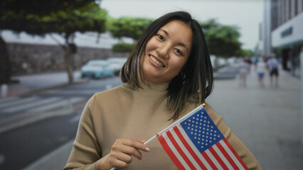 Young chinese woman holding american flag smiling on a city street, symbolizing cultural diversity...