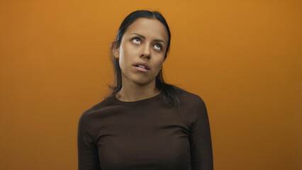 Fototapeta premium Woman expressing boredom over isolated orange wall embodying neutral emotion and casual style featuring young hispanic female in brown sweater with blank background wall setting.