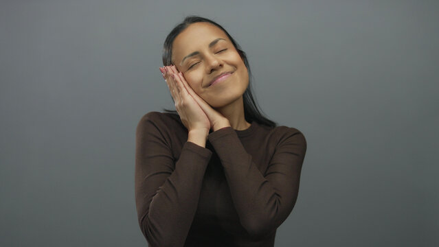 Woman gestures sleep against a grey background, embodying relaxation and tranquility, perfect for concepts of rest or peacefulness in a clean minimal setting.